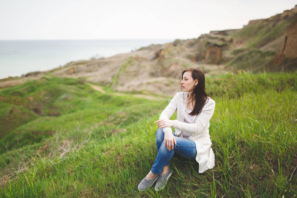 Young brunette concerned beautiful woman in light casual clothes looking aside sitting on grass with crossed hands near water on green field background. Beautiful landscape. Lifestyle leisure concept - Фото, изображение