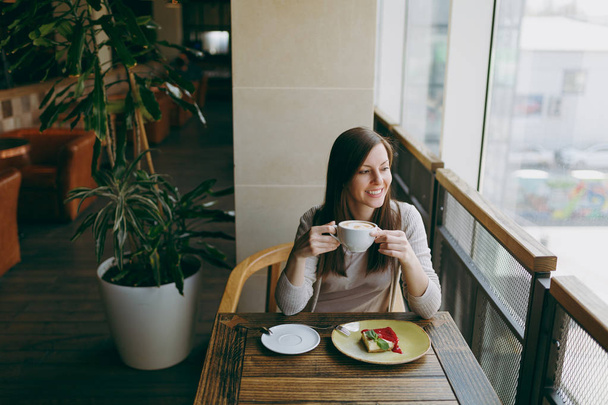 Attractive young woman sitting alone near big window in coffee shop at table with cup of cappuccino, cake, relaxing in restaurant during free time. Young female having rest in cafe. Lifestyle concept - Photo, Image