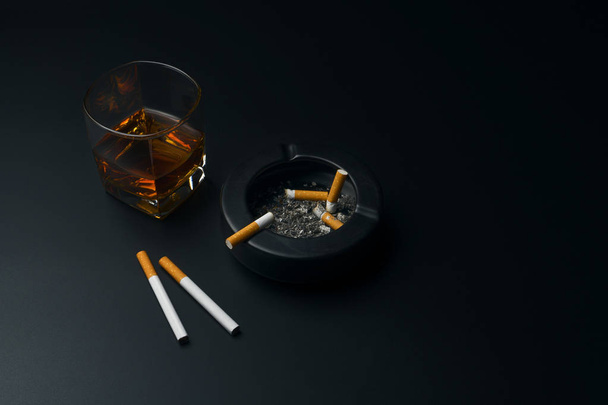 стакан виски бурбон, сигареты и черная керамическая пепельница, полная пепла с окурком, на черном столе
 - Фото, изображение