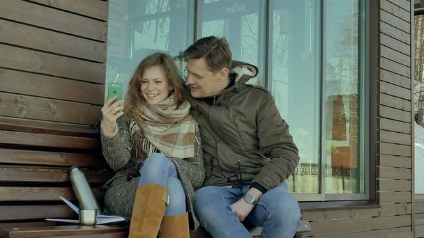 Young woman and man do selfie on the phone and drinking tea from a thermos bottle in a spring park sitting on a bench - Valokuva, kuva