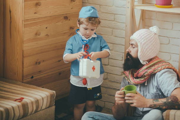 Kid and father play hospital at home, medical education concept. Patient listening to advice of doctor. Bearded man in knitted hat and scarf spending time with his son. - Zdjęcie, obraz