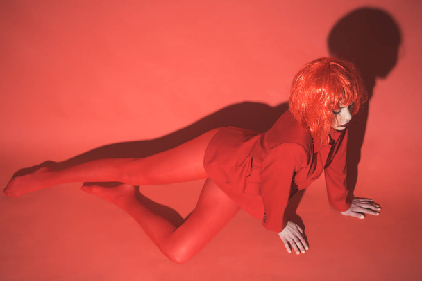 Young female in red outfit and bob style wig isolated on red background. Slim girl with closed eyes posing on floor. Hiding true nature behind fancy appearance, self-finding concept. - 写真・画像