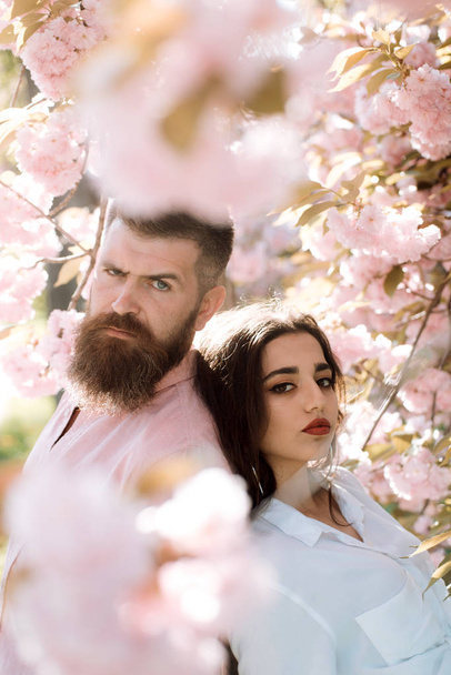Couple in love standing back to back under blooming tree. Bearded man and pretty girl with red lips surrounded by pink petals of cherry blossom. Romantic date in idyllic garden, spring concept. - Φωτογραφία, εικόνα