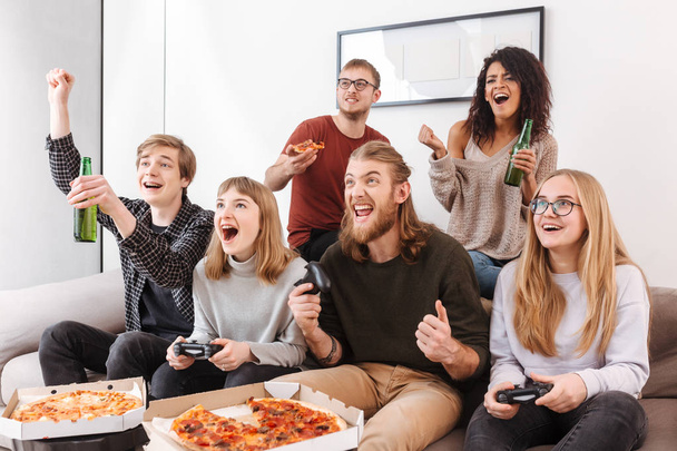 Group of joyful friends sitting on sofa and screaming yeah while spending time together and playing video games. Portrait of young people eating pizza and drinking beer at home - Photo, Image