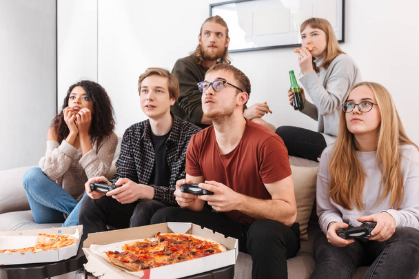 Grupo de amigos sentados en el sofá y mirando cuidadosamente recto mientras pasan tiempo juntos. Retrato de personas jugando videojuegos, comiendo pizza y bebiendo cerveza en casa
 - Foto, imagen