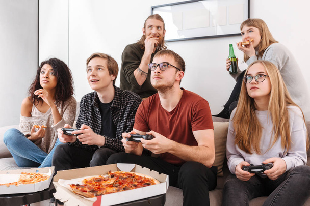 Grupo de amigos reflexivos sentados en el sofá y pasando tiempo juntos mientras juegan videojuegos, comen pizza y beben cerveza en casa
 - Foto, imagen