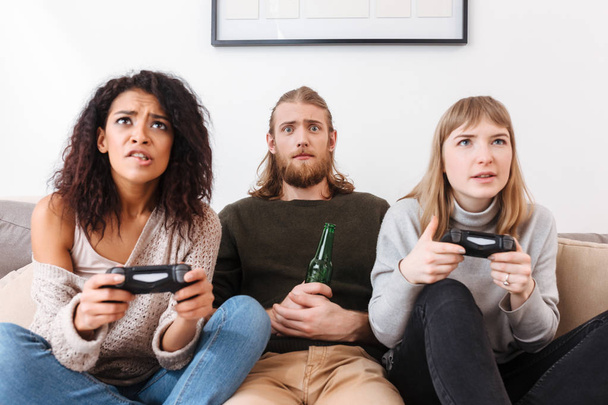 Портрет молодого чоловіка, який сидить з пивом на дивані і дивно дивиться в камеру, поки дві дівчини поблизу грають у відеоігри вдома
 - Фото, зображення