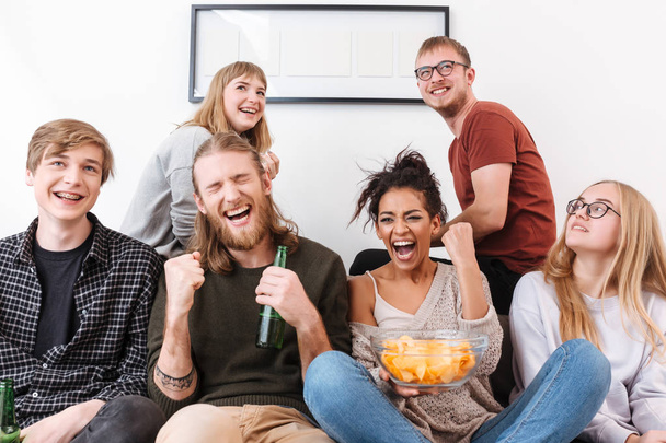 Gruppo di amici gioiosi seduti sul divano a guardare film insieme a patatine e birra a casa
 - Foto, immagini