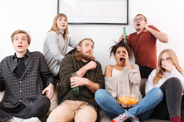 Закрыть фотографию напуганных друзей, сидящих на диване и смотрящих фильм ужасов вместе с чипсами и пивом дома
 - Фото, изображение