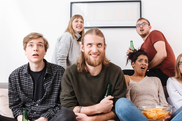 Retrato del joven sonriente y sus amigos asustados sentados en el sofá con papas fritas y cerveza y viendo películas de terror juntos en casa
 - Foto, Imagen
