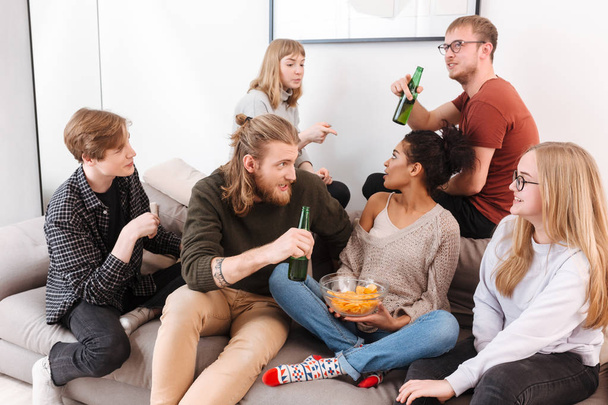 Gruppe junger Freunde sitzt auf dem Sofa und diskutiert mit Chips und Bier zu Hause - Foto, Bild