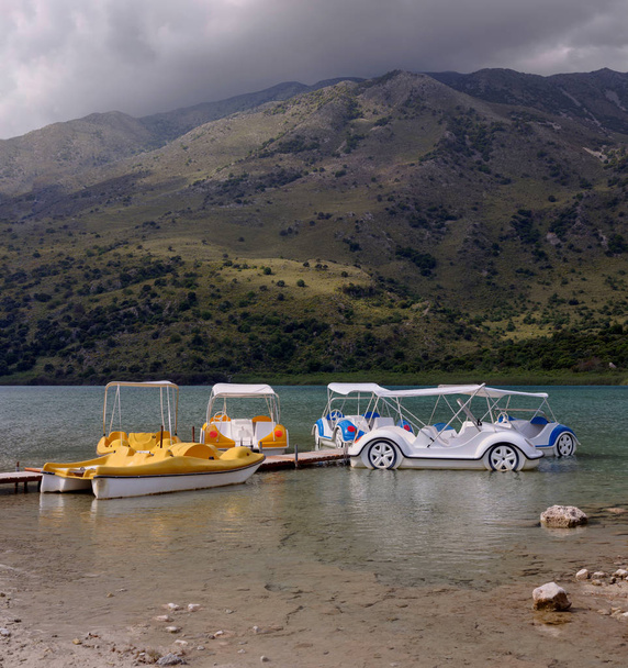 Вид на природное озеро Кофас, прогулочные лодки и горы вокруг в пасмурный весенний день (остров Крит, Греция)
 - Фото, изображение
