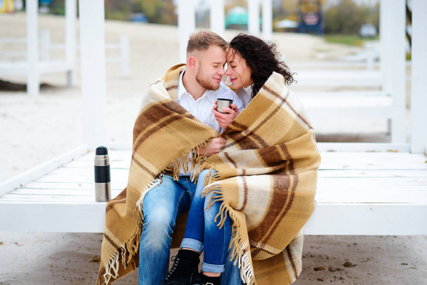 junges Paar umarmt und in eine warme Decke gehüllt sitzt am Herbststrand und trinkt Kaffee aus einer Thermoskanne - Foto, Bild