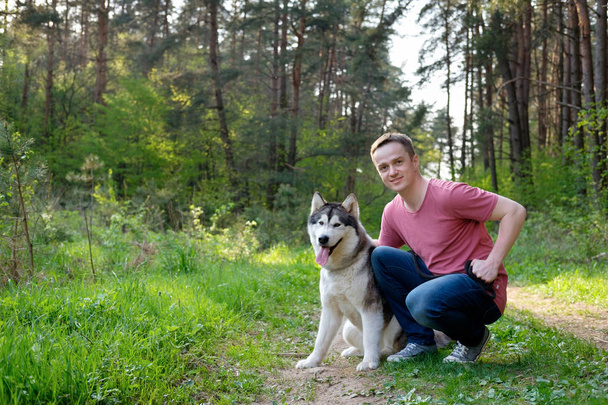 attractive young man in a pink T-shirt and blue jeans with his dog malamute on a walk in the forest - Φωτογραφία, εικόνα