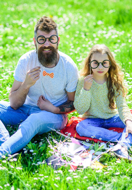 Dad and daughter sits on grass at grassplot, green background. Child and father posing with eyeglases photo booth attributes at meadow. Family spend leisure outdoors. Smart and clever concept - Photo, image