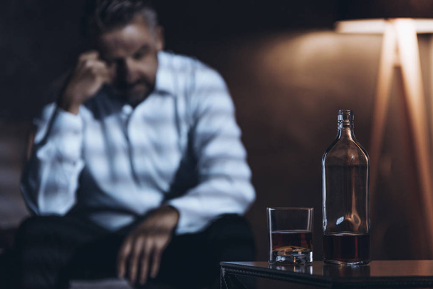Stressed man sitting in a dark room with a bottle and glass with alcohol - Foto, Imagen