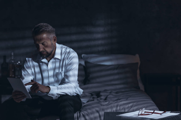 Exhausted middle-aged male sitting on a bed in a hotel room at night and looking into a notepad during his corporate business trip - Fotoğraf, Görsel