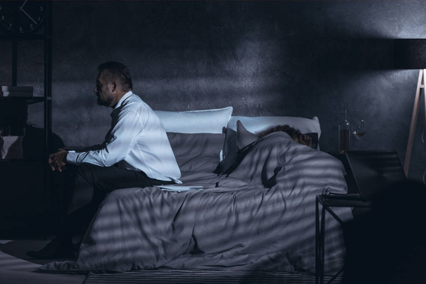 Dark gray room with a side view of a mature man sitting on a bed and having second thoughts after cheating and a woman sleeping under the cover - Fotografie, Obrázek