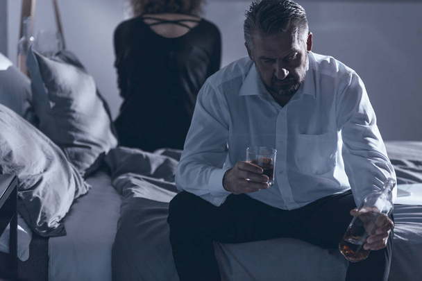 Husband sitting on the bed and holding alcohol while having a fight with his wife - Foto, Imagen