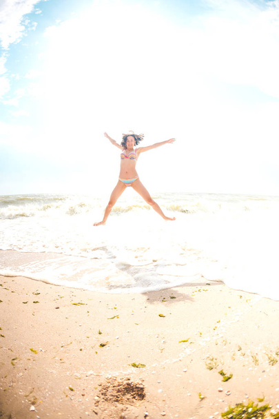 Une fille saute sur la plage et place ses mains larges. Une femme mince repose sur la côte de la mer. Brunette au bord de l'océan. Fitness dans la nature
. - Photo, image