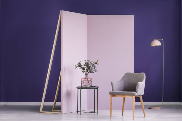 Real photo of twigs in a pink, glass vase on a metal table next to a gray armchair in cozy, pastel interior - Photo, Image