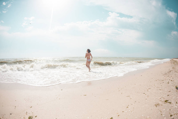 Tyttö kävelee pitkin rantaa. Nainen seisoo meren hiekkarannalla. Brunette uimapuvussa nauttii merestä
. - Valokuva, kuva