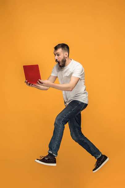 Image of young man over orange background using laptop computer while jumping. - Foto, imagen
