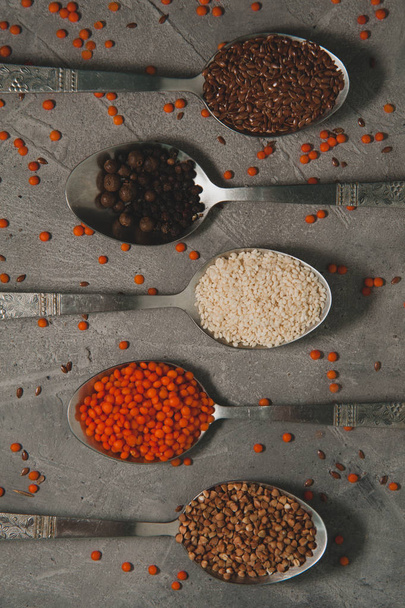 Spoons with different superfoods - flax seeds, sesame, pepper, red lentils, buckwheat on a gray background. Vertical photo - Foto, Imagem