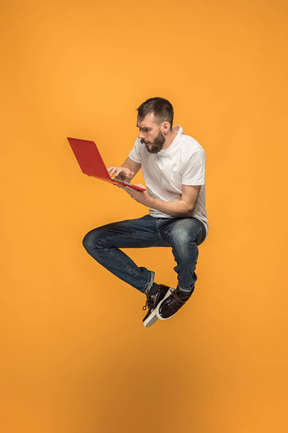 Image of young man over orange background using laptop computer while jumping. - 写真・画像