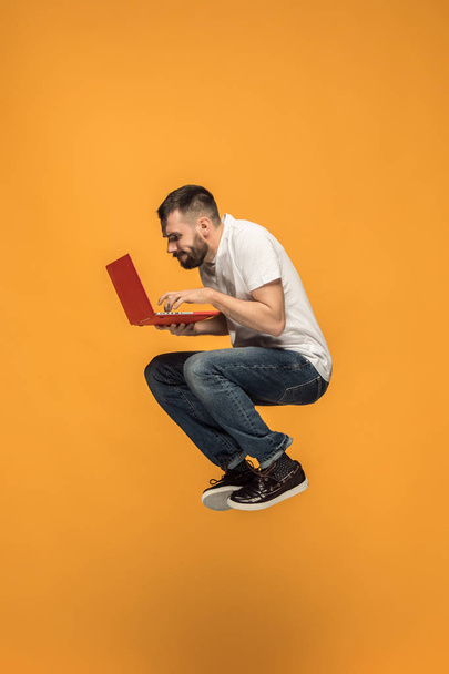 Image of young man over orange background using laptop computer while jumping. - Foto, immagini