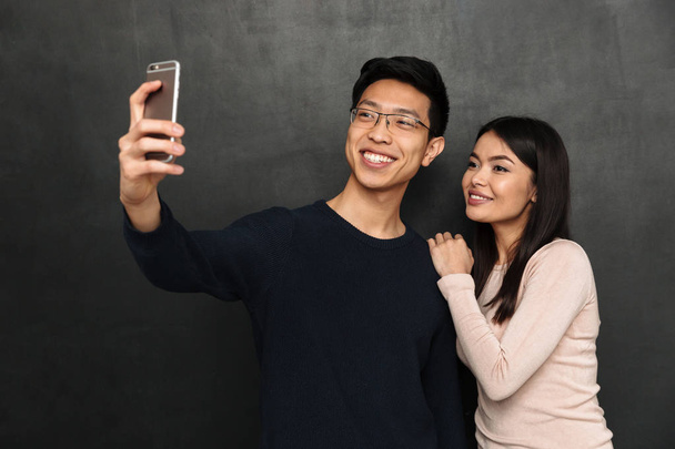 Cheerful asian couple posing together and making selfie on smartphone over black background - Photo, image