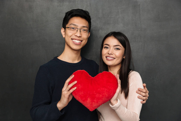 Happy asian couple posing together while holding heart toy and looking at the camera over black background - Zdjęcie, obraz