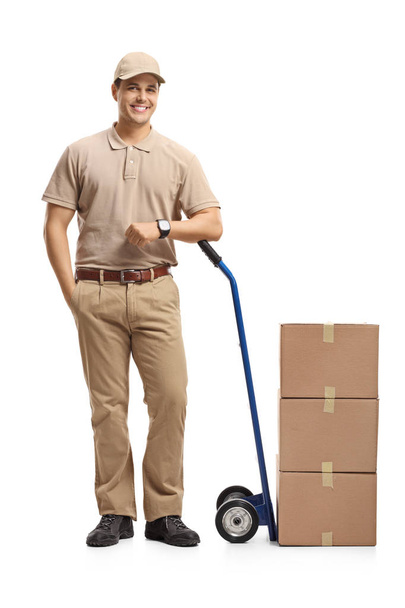 Full length portrait of a delivery man leaning on a hand truck loaded with boxes isolated on white background - Фото, изображение