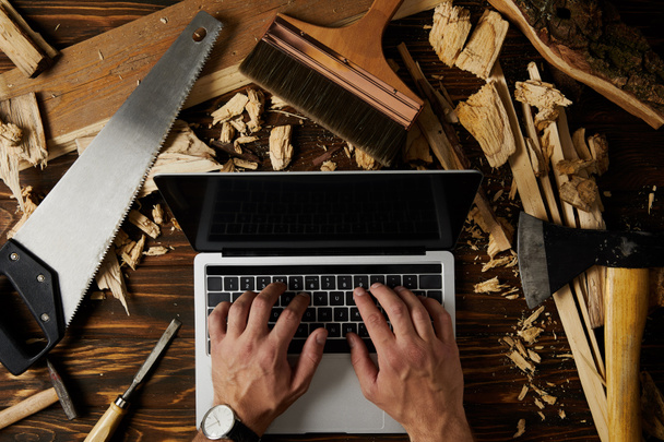 обрезанное изображение плотника, печатающего на ноутбуке в окружении различных инструментов на столе
  - Фото, изображение