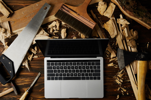 ноутбук с чистым экраном, окруженный топором, ручной пилой, зубилом, кистью и молотком на деревянном столе
 - Фото, изображение