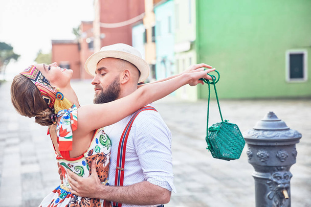 Romantic couple on colorful Burano island streets. Woman in colorful dress and headband with little straw bag, man in white shirt, straw hat and dark pants with suspenders  - Photo, image