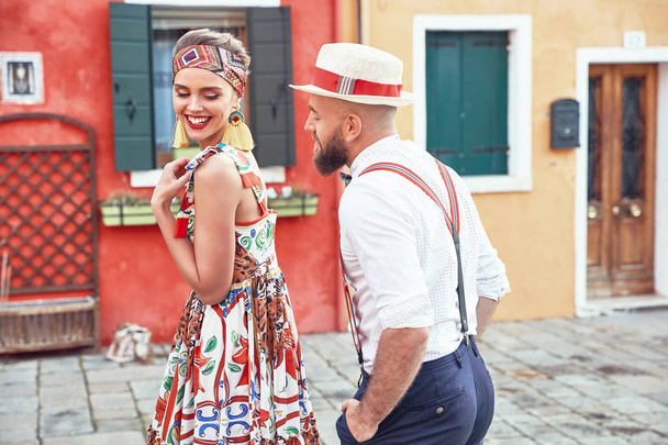 Romantic couple having fun on colorful Burano island streets. Woman in colorful dress and headband with little straw bag, man in white shirt, straw hat and dark pants with suspenders  - Fotó, kép