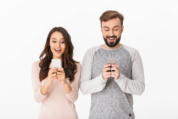 Image of excited young loving couple standing isolated over white background. Looking aside using mobile phones. - 写真・画像