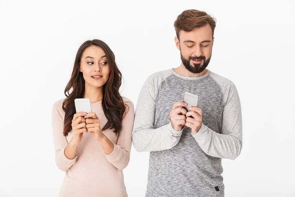 Image of excited young loving couple standing isolated over white background. Looking aside using mobile phones. - Фото, изображение
