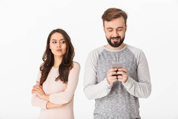 Image of displeased young woman looking at her man standing isolated over white background using mobile phone. - Photo, Image
