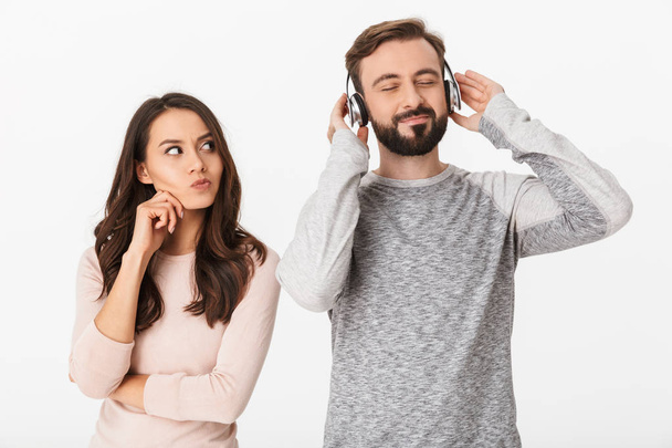 Imagen de una joven disgustada mirando a su hombre de pie aislado sobre fondo blanco escuchando música con auriculares
. - Foto, imagen