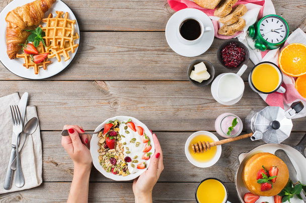 Essen und Trinken, gesundes Morgenessen. Frühstück mit Pfannkuchen, Waffeln, Croissant-Sandwich und Müsli auf dem Holztisch. Flachbild-Hintergrund mit Frauenhänden - Foto, Bild