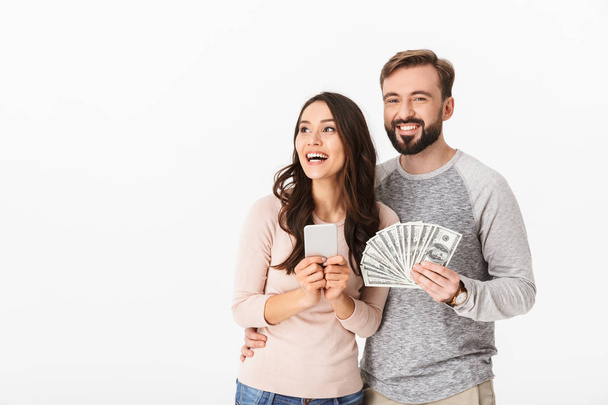 Photo of happy young loving couple standing isolated over white background holding money using mobile phone. - Foto, imagen