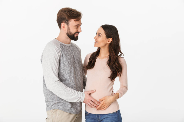 Photo of cheerful young man standing with his pregnant woman isolated over white background wall. Looking at each other - Photo, Image
