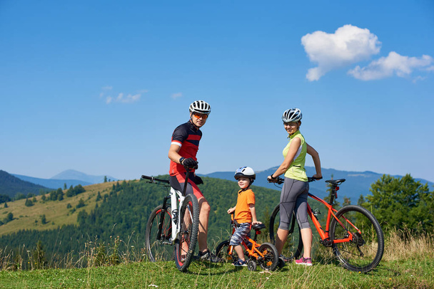 Young modern family tourists resting on bikes on grassy hill with distant mountains view background, Active lifestyle, traveling and happy relations concept  - Photo, Image