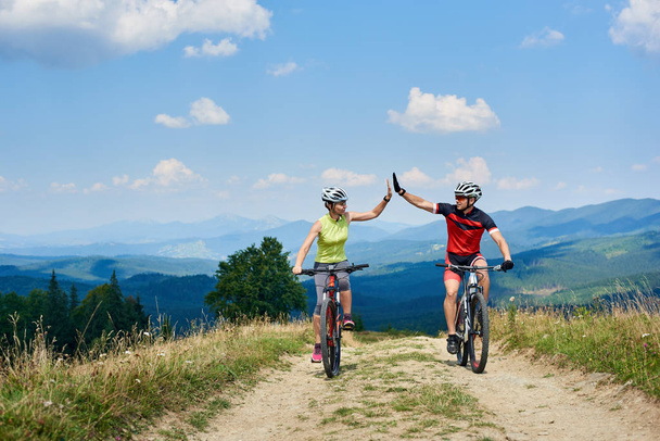 Aktive Paarradler in professioneller Sportbekleidung und Helm, die sich gegenseitig hohe fünf geben, während sie im Sommer auf der Bergstraße Cross-Country-Bikes fahren, aktiver Lebensstil und Outdoor-Sportkonzept - Foto, Bild