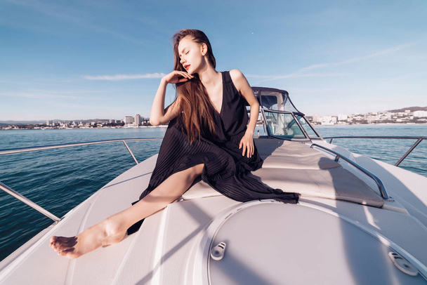 long-haired young girl in black summer clothes sits on her white yacht, posing in the sun and enjoying the warmth - Foto, Imagem