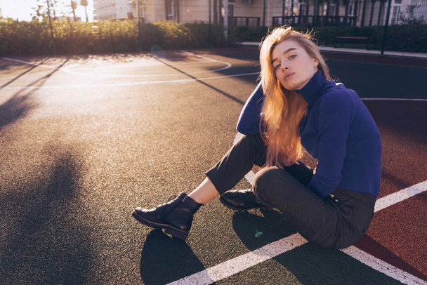 stylish blond model girl in blue sweater sits on playground and posing in morning sun rays - Фото, изображение