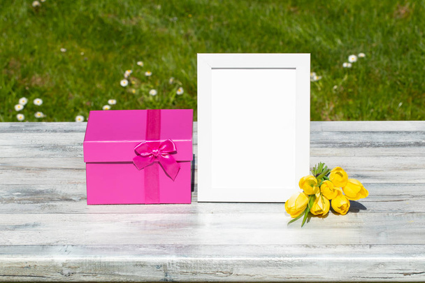 Gift box, flowers and white vintage frame with place for your text on rustic bright wooden table. Green natural background. Card concept. Outdoor. - Foto, imagen