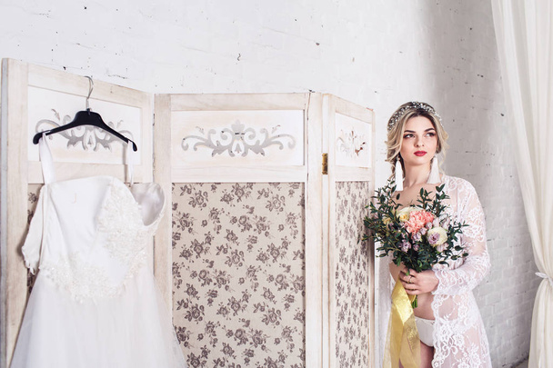 Braut, in Peignoir und Unterwäsche gekleidet, steht neben Schild mit Brautkleid und hält Strauß in ihren Händen - Foto, Bild